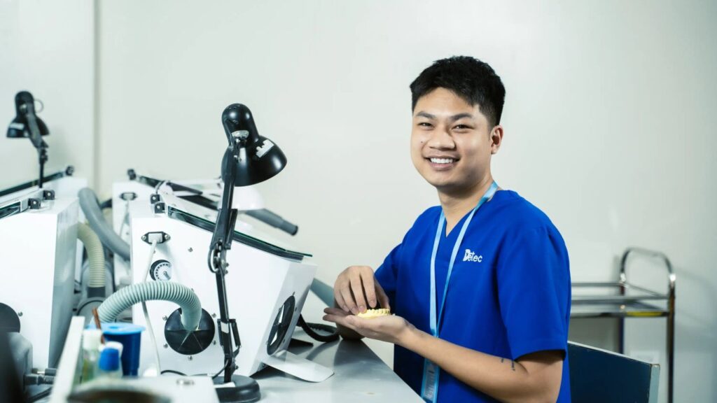 A person in a blue uniform works with dental prosthetics in a lab.