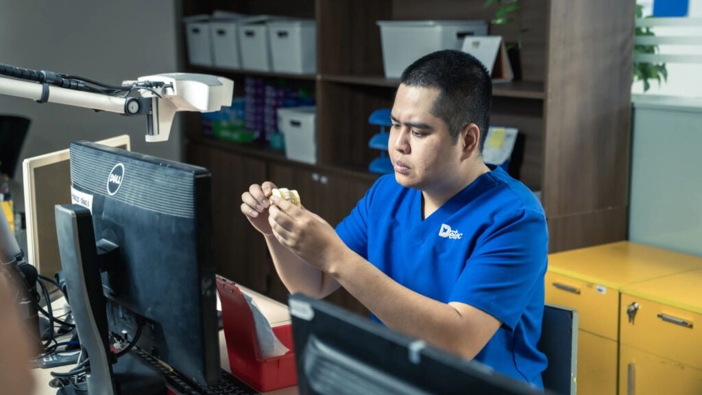 A person in a blue uniform examines a dental mold at a desk.